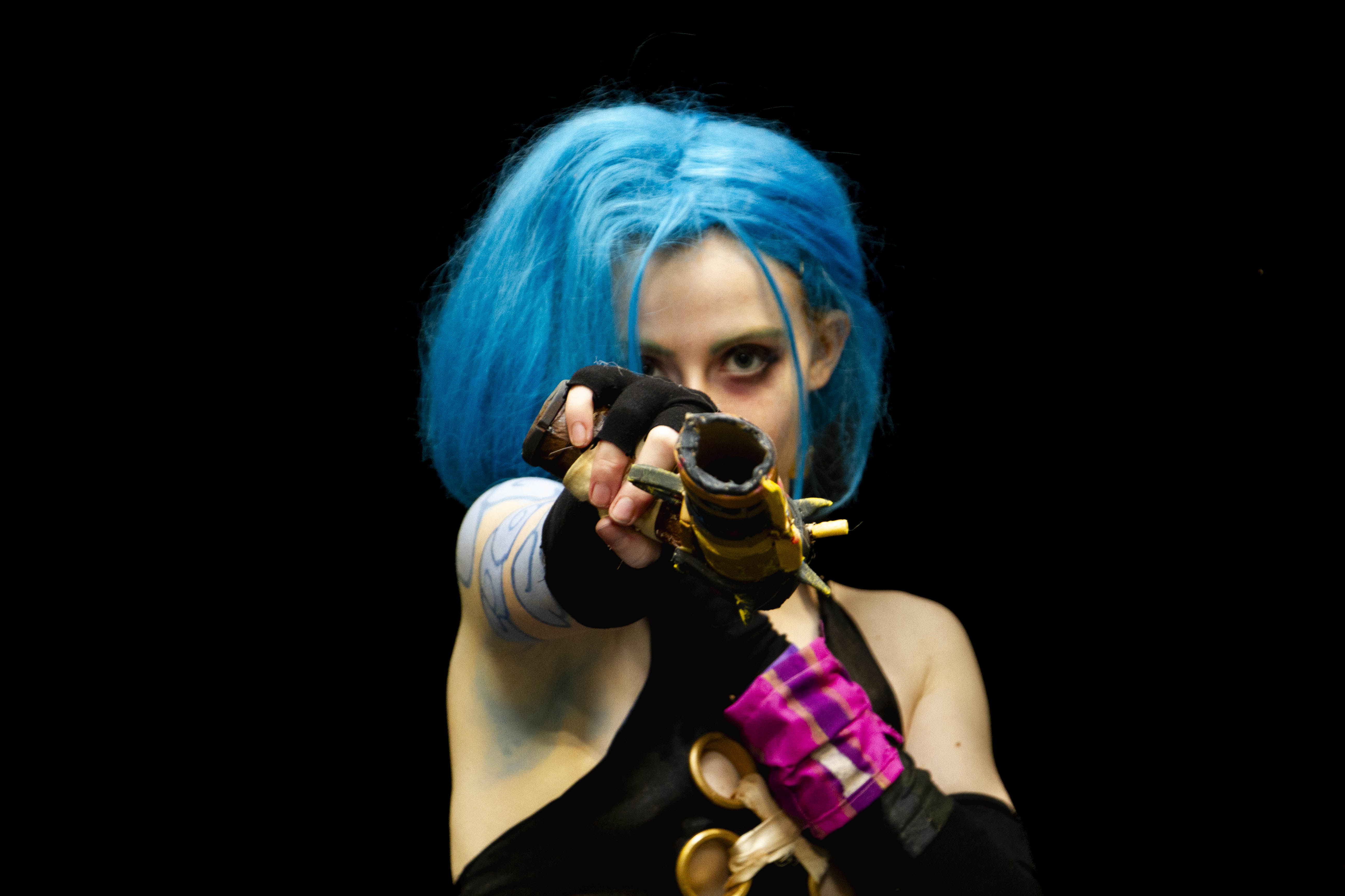 A person cosplaying Jinx from the TV Show Arcane. She is pointing a gun at the camera, with a dark expression.