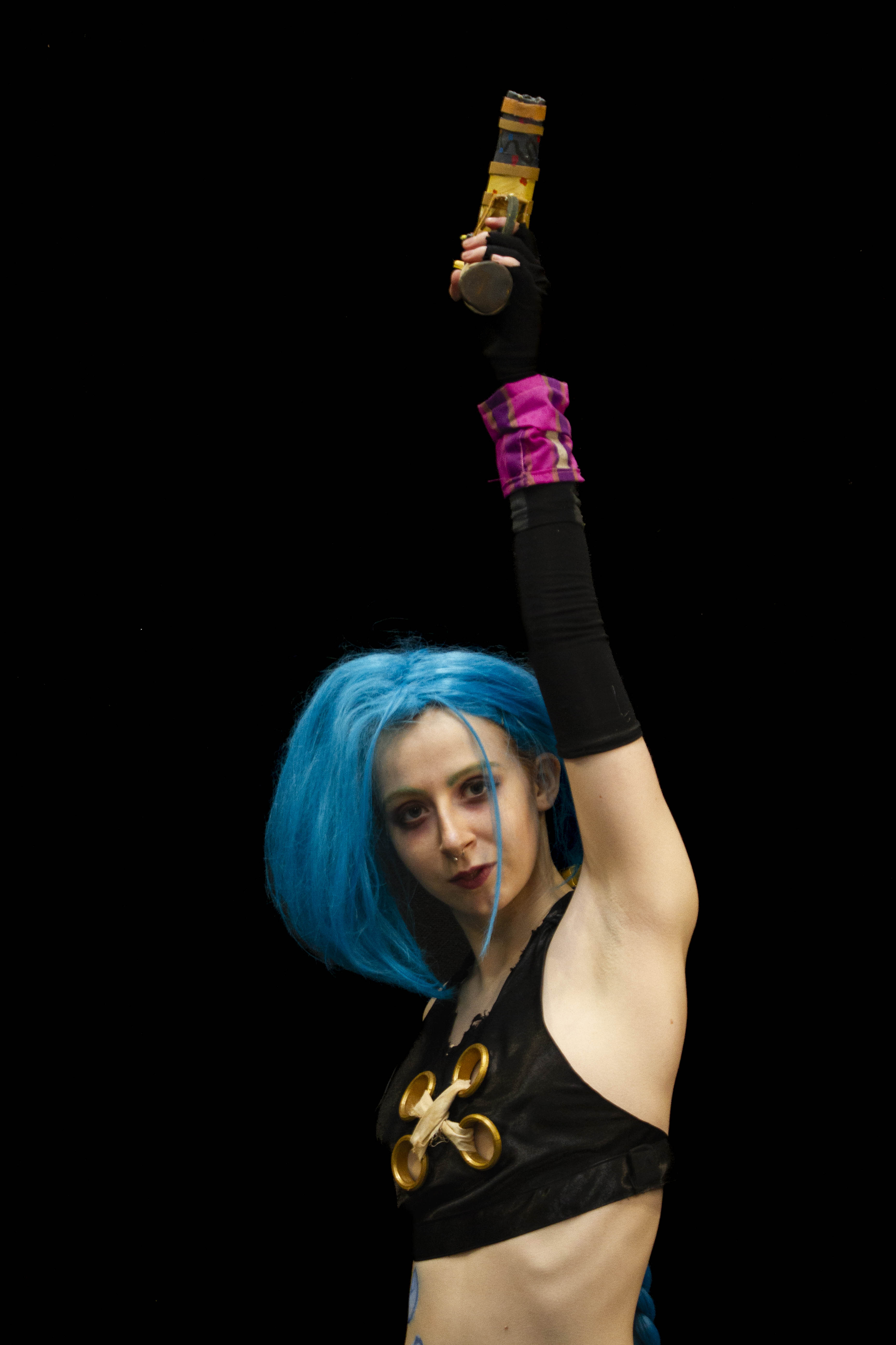 A person cosplaying Jinx from the TV Show Arcane. She is pointing a gun upwards, looking at the camera with a neutral expression.