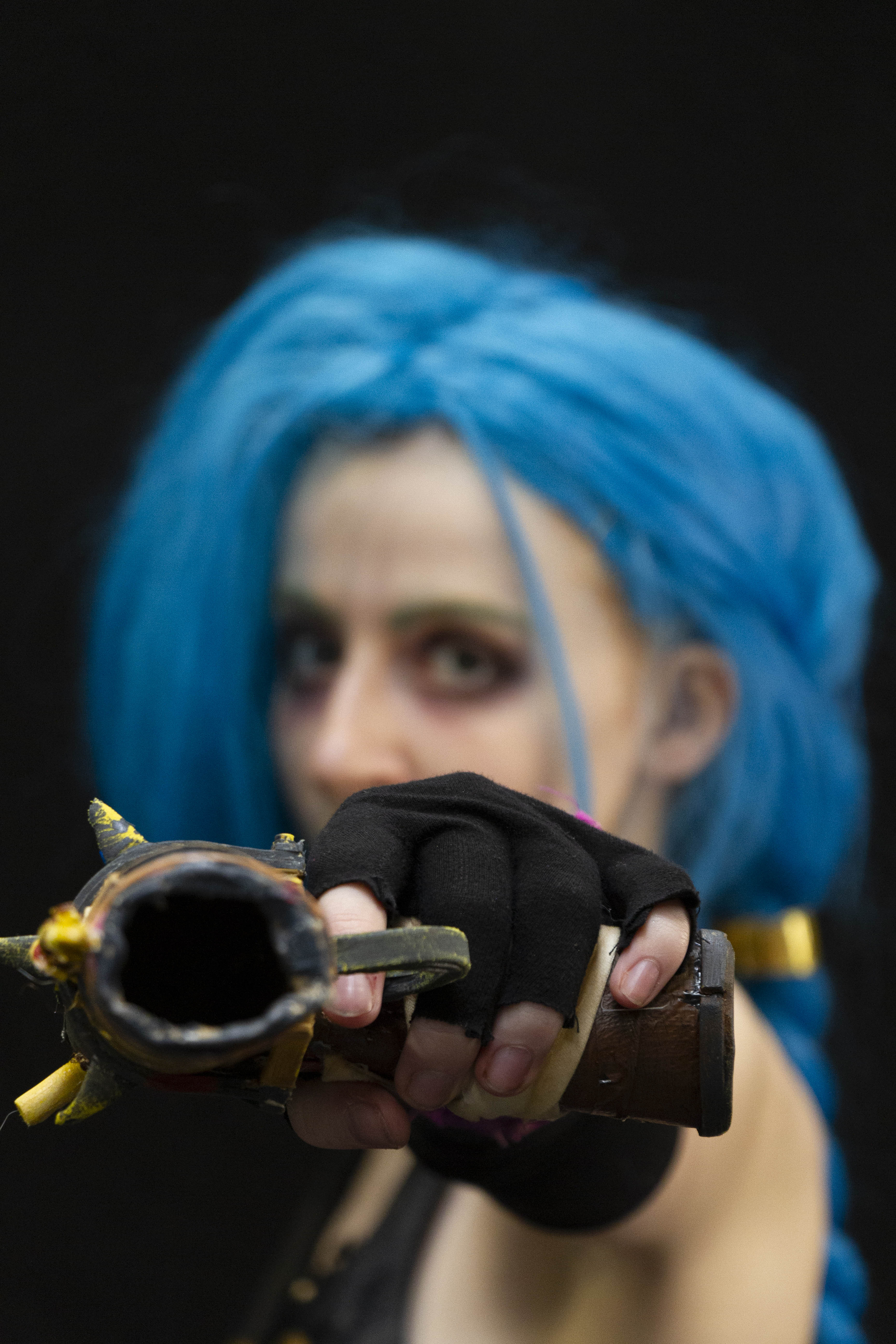 A gun, held by a person cosplaying Jinx from the TV Show Arcane. She is out of focus, pointing the gun at the camera, looking at the camera with a neutral expression.